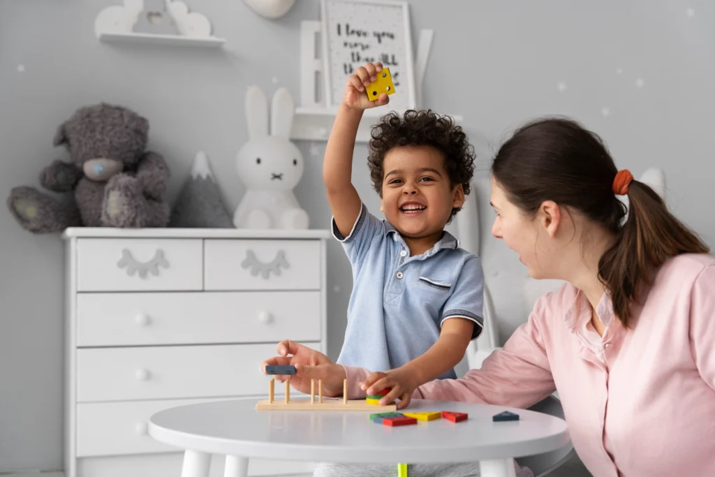 A smiling child raises a yellow block while playing an educational stacking game with a caregiver at a white table in a cozy, decorated room.