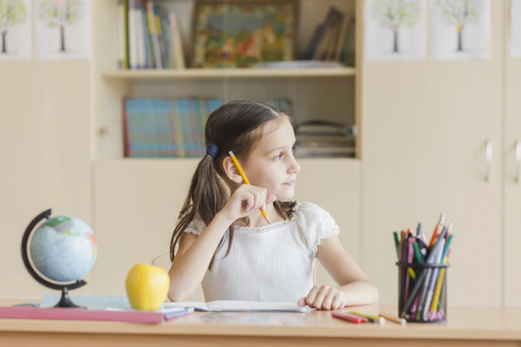 Child sitting at a classroom desk thinking while holding a pencil, surrounded by school supplies and a globe.