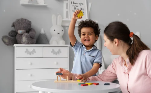 A smiling child raises a yellow block while playing an educational stacking game with a caregiver at a white table in a cozy, decorated room.